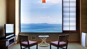 A room with a large window offering a view of the sea, two chairs, a small table, and a television.