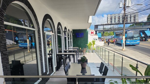 Een terras met tafels en stoelen, uitzicht op de straat, en een gebouw met een 'H'-bordje dat een ziekenhuis in de buurt aanduidt.