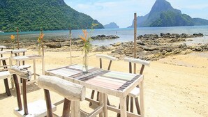 Een restaurant aan het strand met houten tafels en stoelen, een vaas met bloemen en uitzicht op zee en bergen.