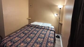 A small, single-bed room with a floral-patterned bedspread, a bedside lamp, and beige walls.