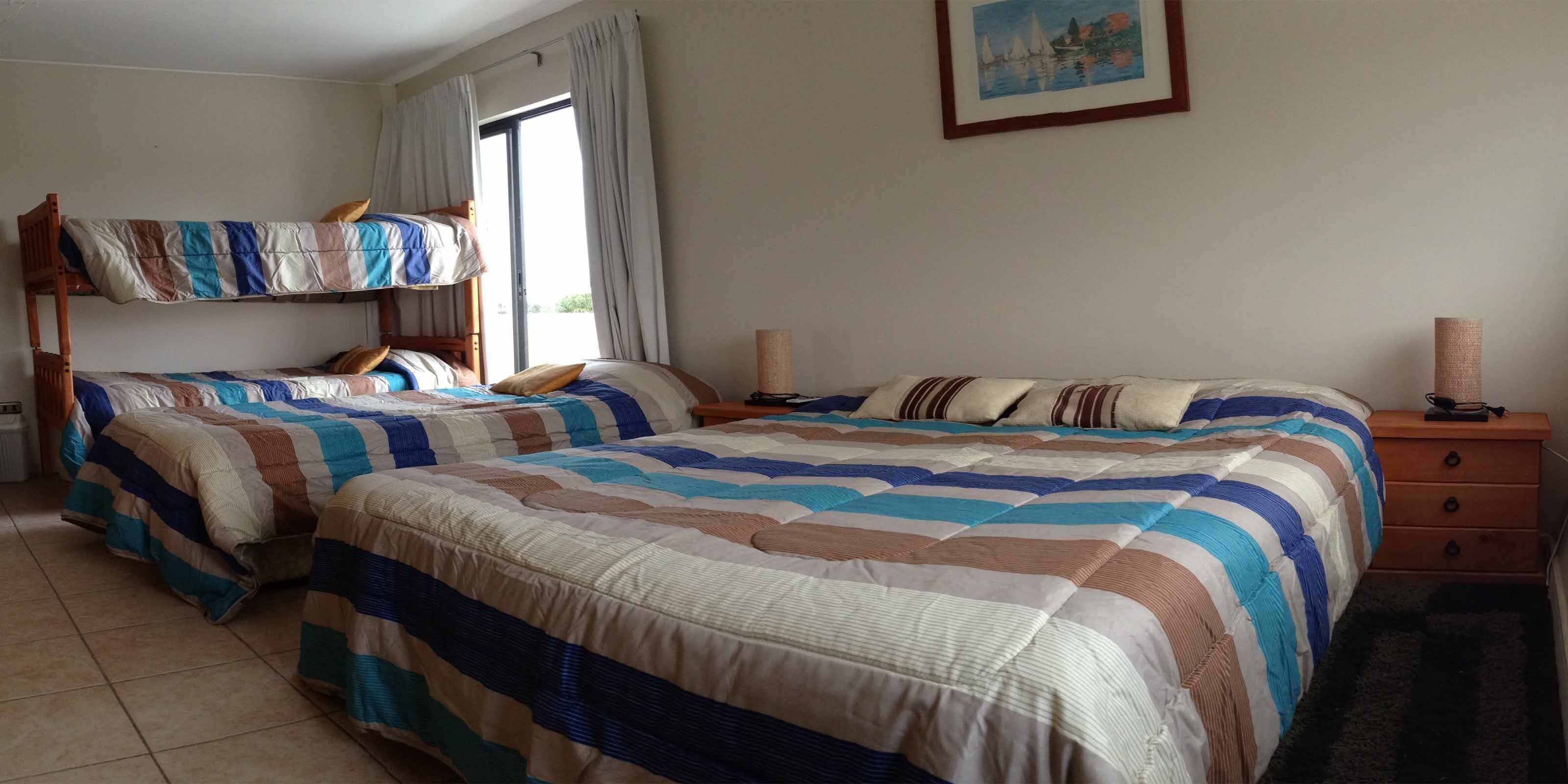 Une chambre avec deux lits superposés, une table de chevet en bois avec une lampe, un tableau encadré au mur et une fenêtre avec des rideaux.