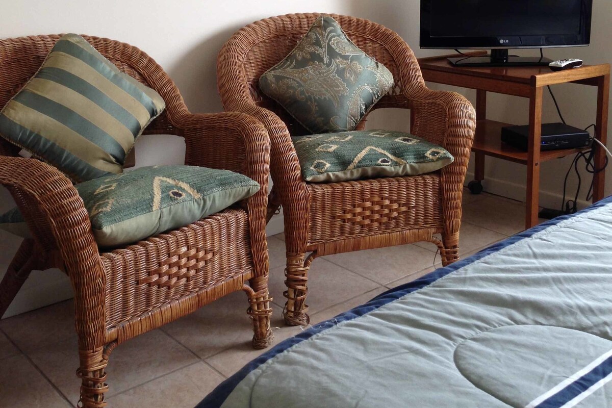 A room with two wicker chairs, a bed, a television on a wooden stand, and a framed picture on the wall.