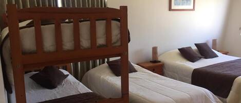 A room with two single beds, a wooden bunk bed, a bedside table with a lamp, and a framed picture on the wall.