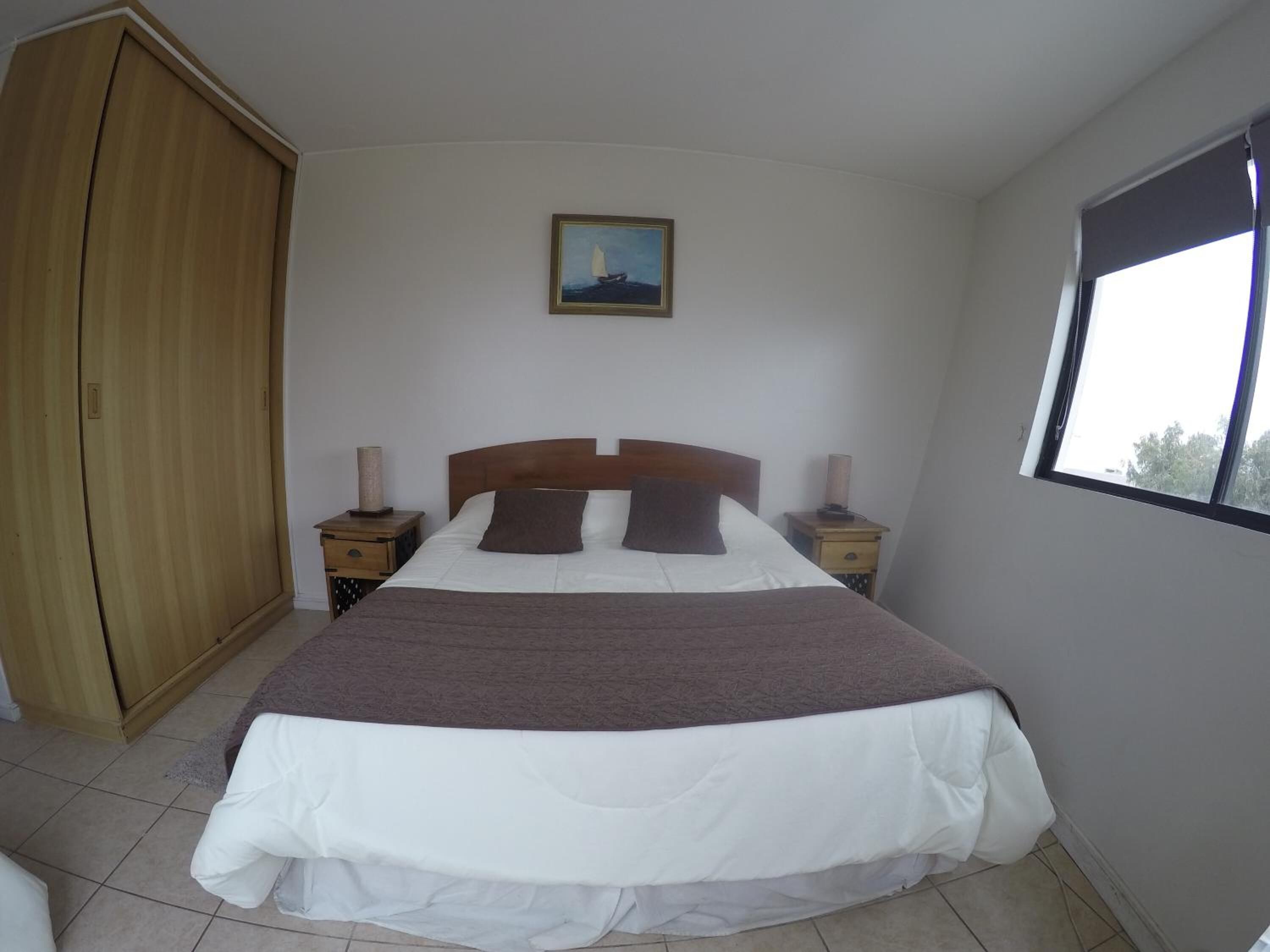A bedroom with a bed, a wardrobe, a nightstand, a lamp, a painting, and a window.