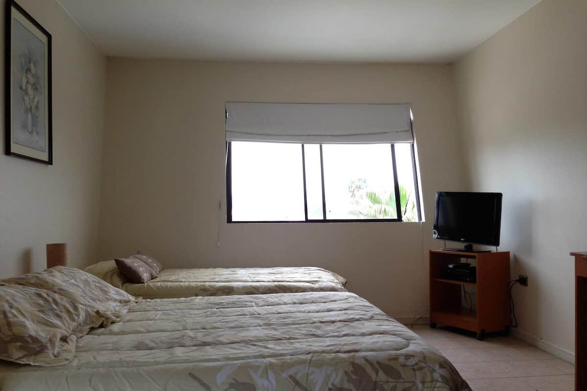 A bedroom with two beds, a television on a wooden stand, and a window with a white blind.