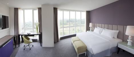 Habitación de hotel con una cama grande, un escritorio y una silla. Se ve follaje verde a través de la ventana.