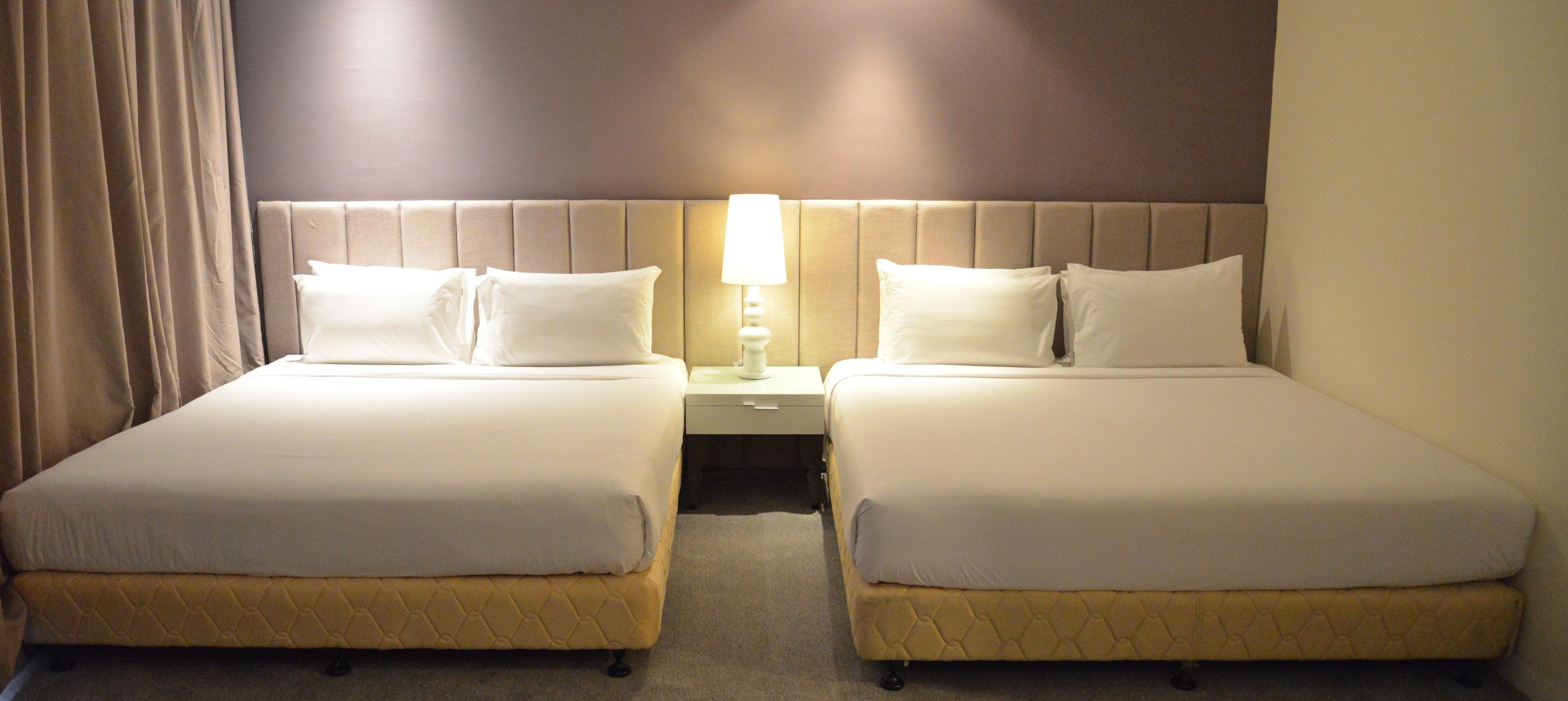 A hotel room with two beds, a nightstand with a lamp, and a curtain.