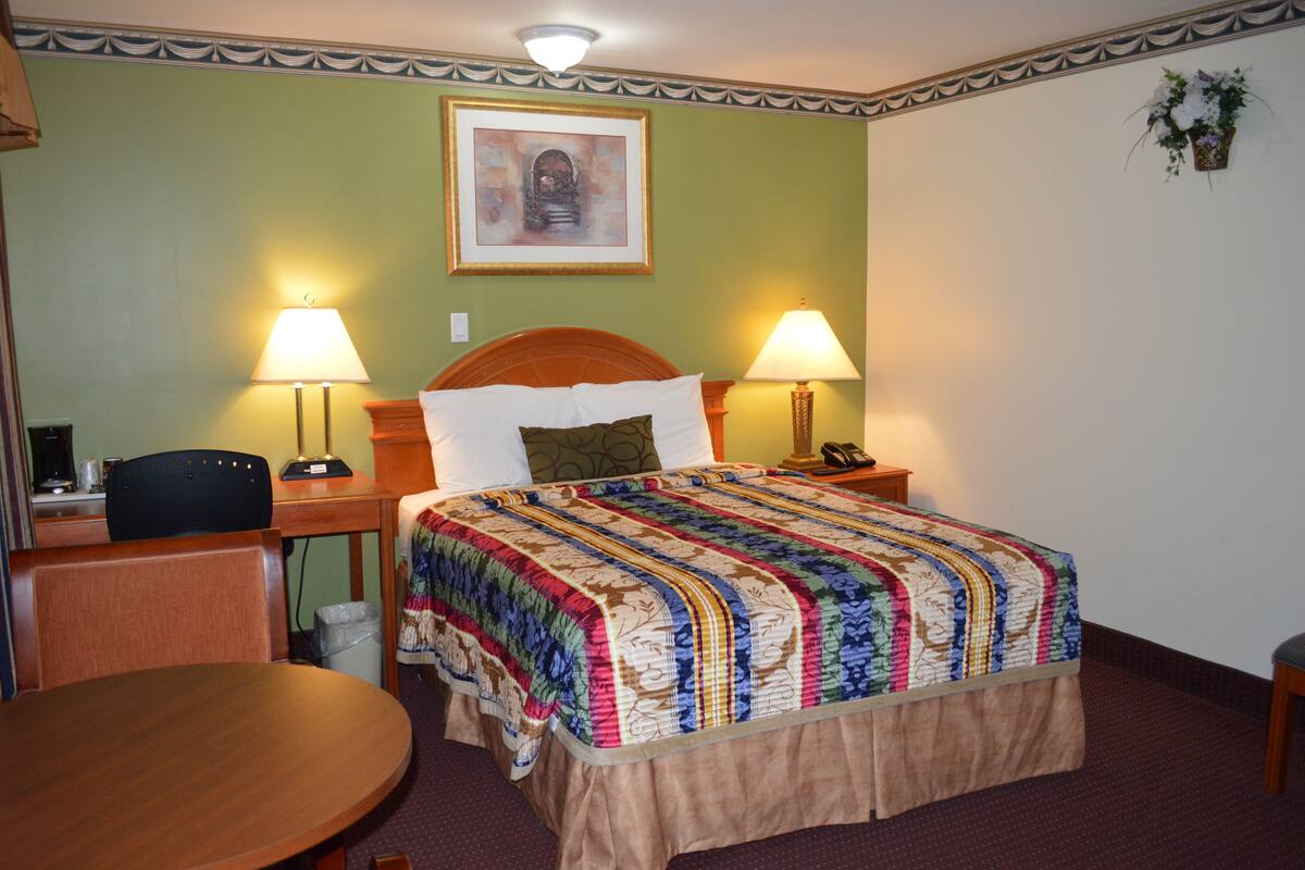 A hotel room with a bed, two bedside lamps, a desk with a chair, a small table, and a framed picture on the wall.