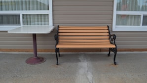 Un banc en bois doté d’accoudoirs en métal et une table blanche devant un bâtiment dont le revêtement extérieur est composé de panneaux horizontaux et qui possède une fenêtre avec des stores verticaux.