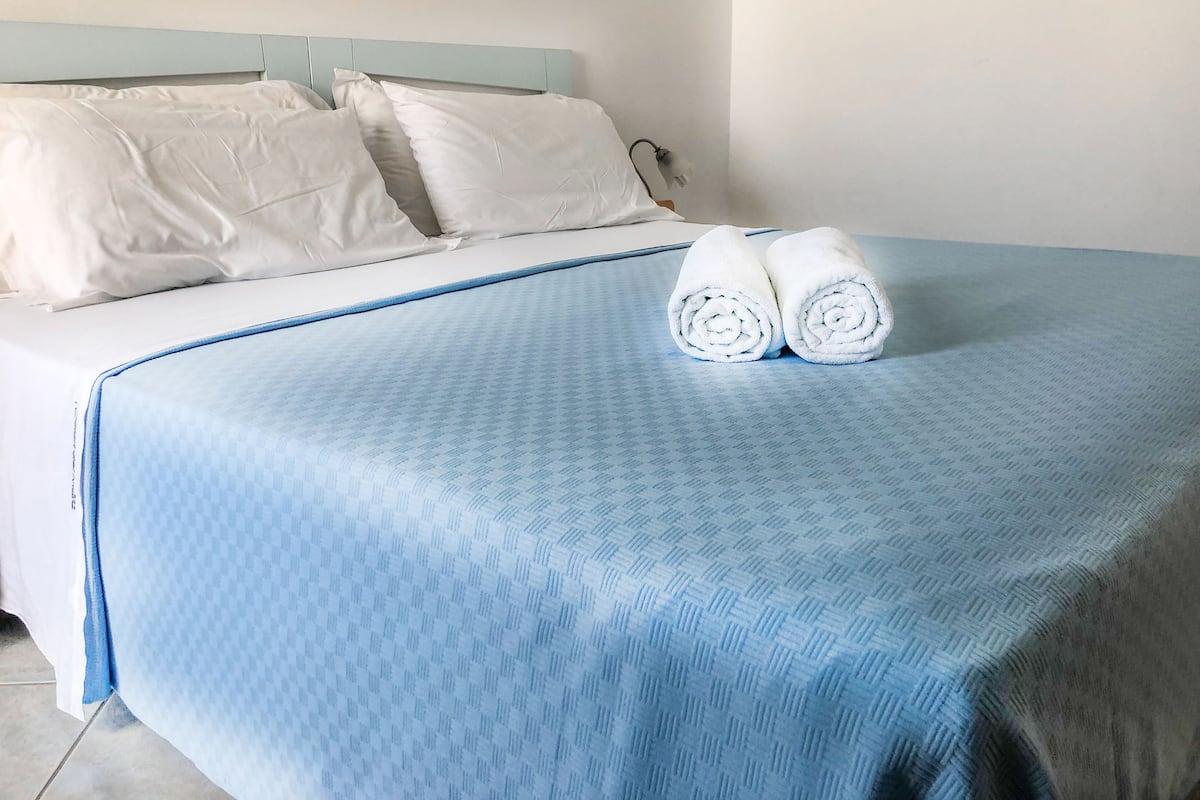 Un lit bien fait, avec des oreillers blancs et une couvre-lit bleue, deux serviettes blanches roulées posées sur le lit, et une tête de lit bleu clair.