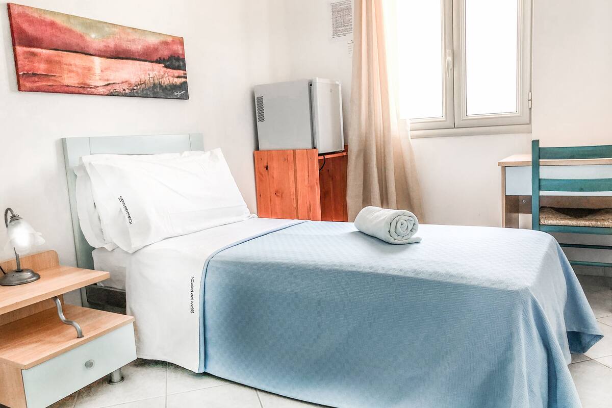 Un lit bien fait, recouvert d’une couverture bleue, avec un oreiller blanc et une serviette enroulée posée sur le lit. On aperçoit une table de chevet en bois avec une lampe, une chaise en bois et une petite table également équipée d’une lampe. Un tableau est accroché au mur, au-dessus du lit, et une fenêtre avec des rideaux est visible.