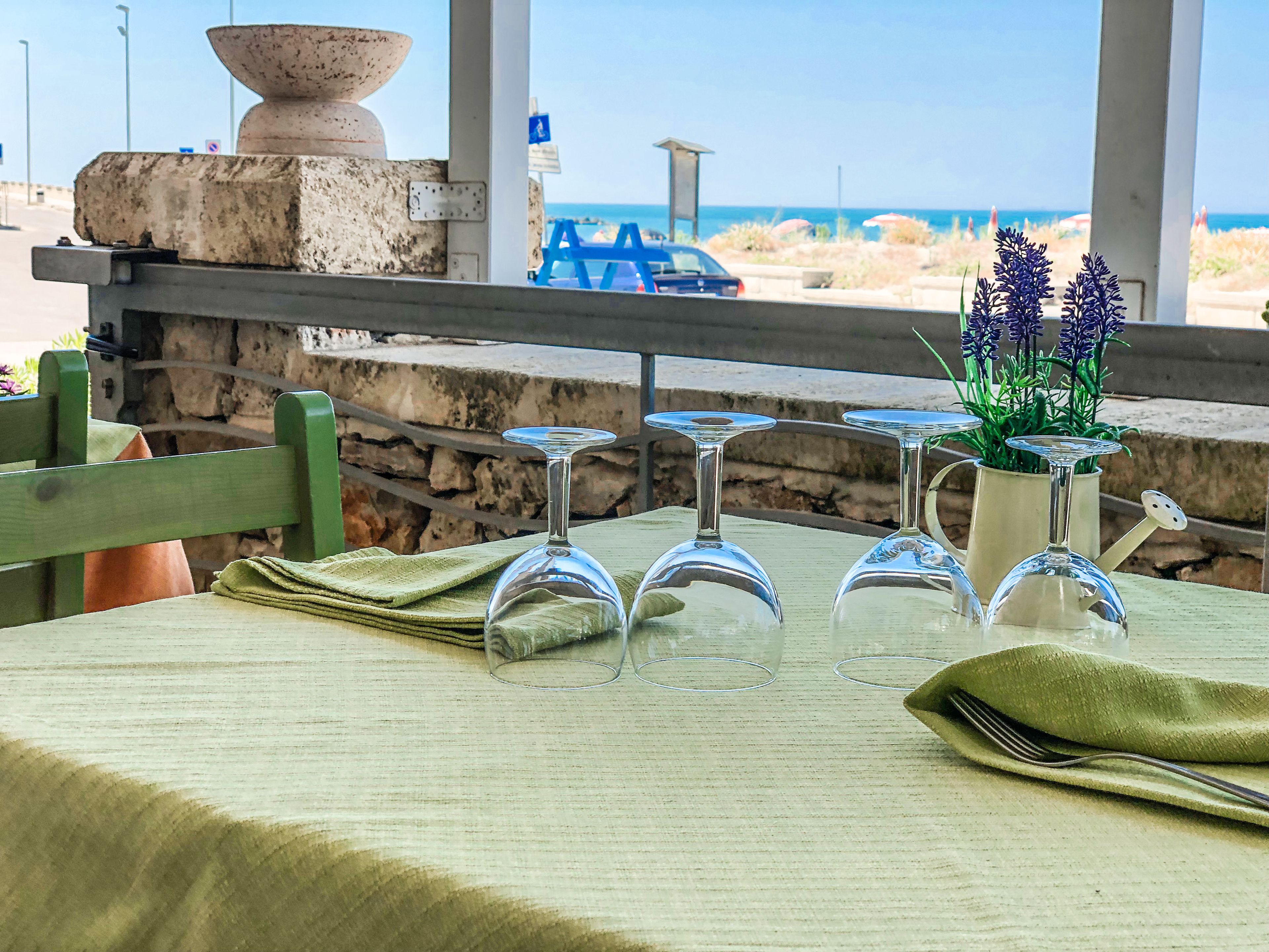 A table set with wine glasses and a fork, overlooking a beach with a blue car and a stone structure in the background.