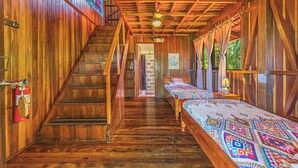 Interior de una cabaña de madera con una escalera, una cama con una manta colorida y un ventilador de techo.