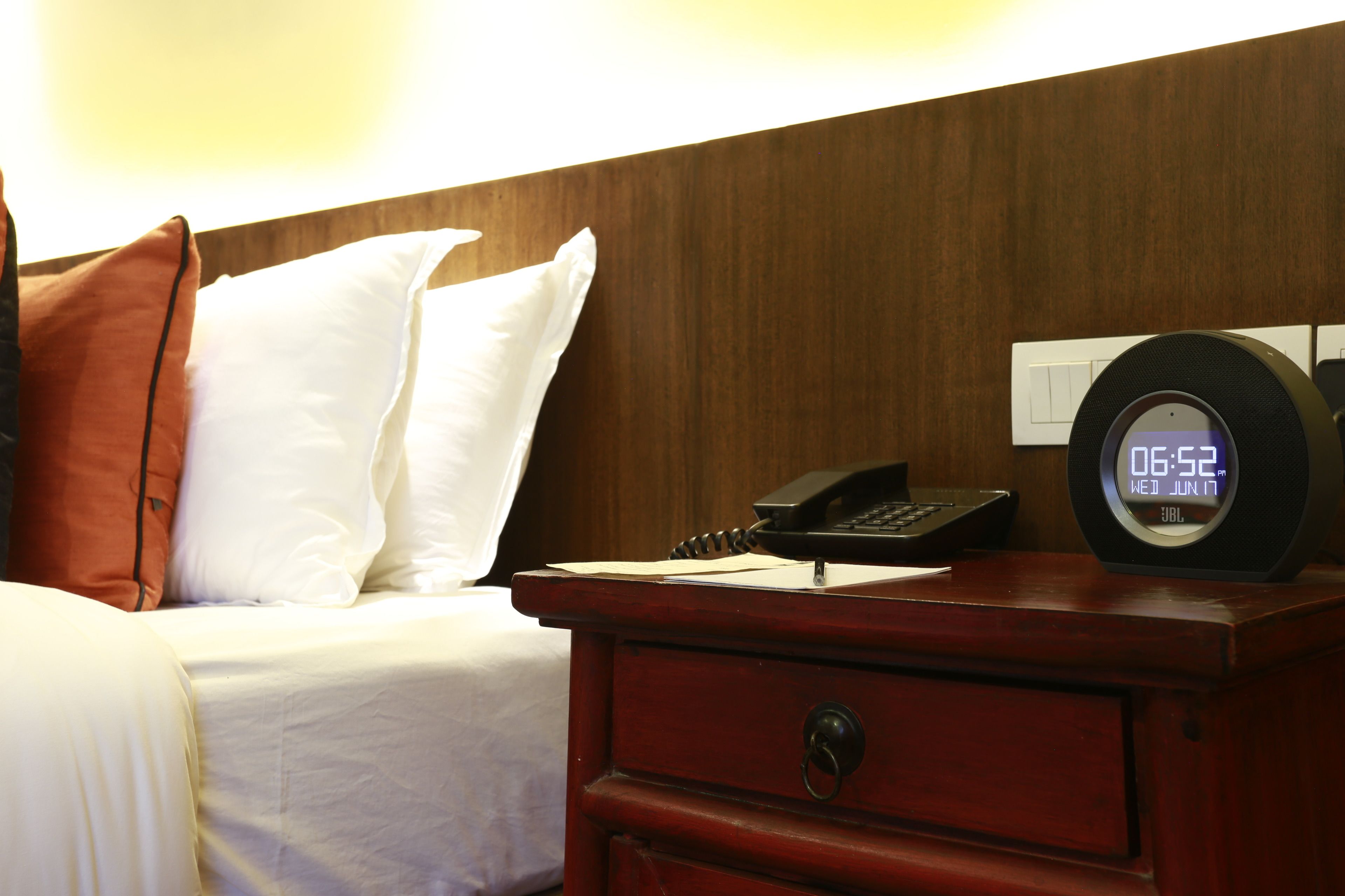 Habitación de hotel con cama, almohadas, mesita de noche de madera, teléfono y reloj digital que muestra la hora.