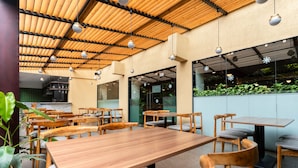 A restaurant with wooden tables and chairs, a high wooden ceiling with hanging lights, and a view of greenery outside. - Hotel Shambala (Kathmandu)