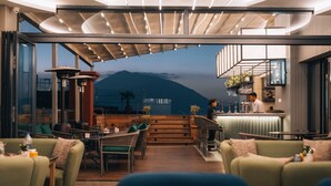 A modern outdoor seating area with a view of mountains, furnished with green sofas and tables, and a bar area with a bartender. - Hotel Shambala (Kathmandu)