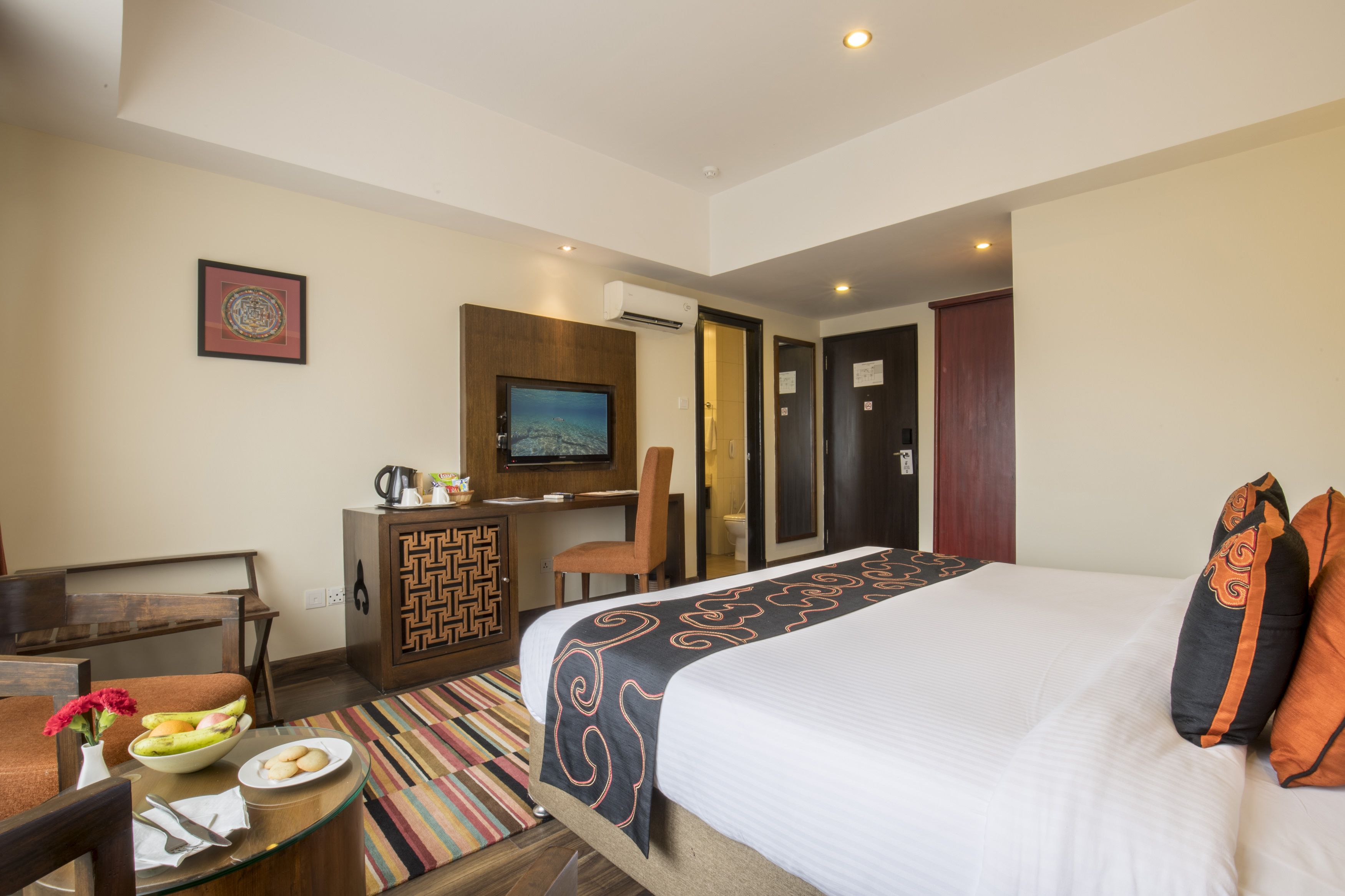 Habitación de hotel con cama, escritorio, silla, televisor y un pequeño rincón de comedor con un frutero.