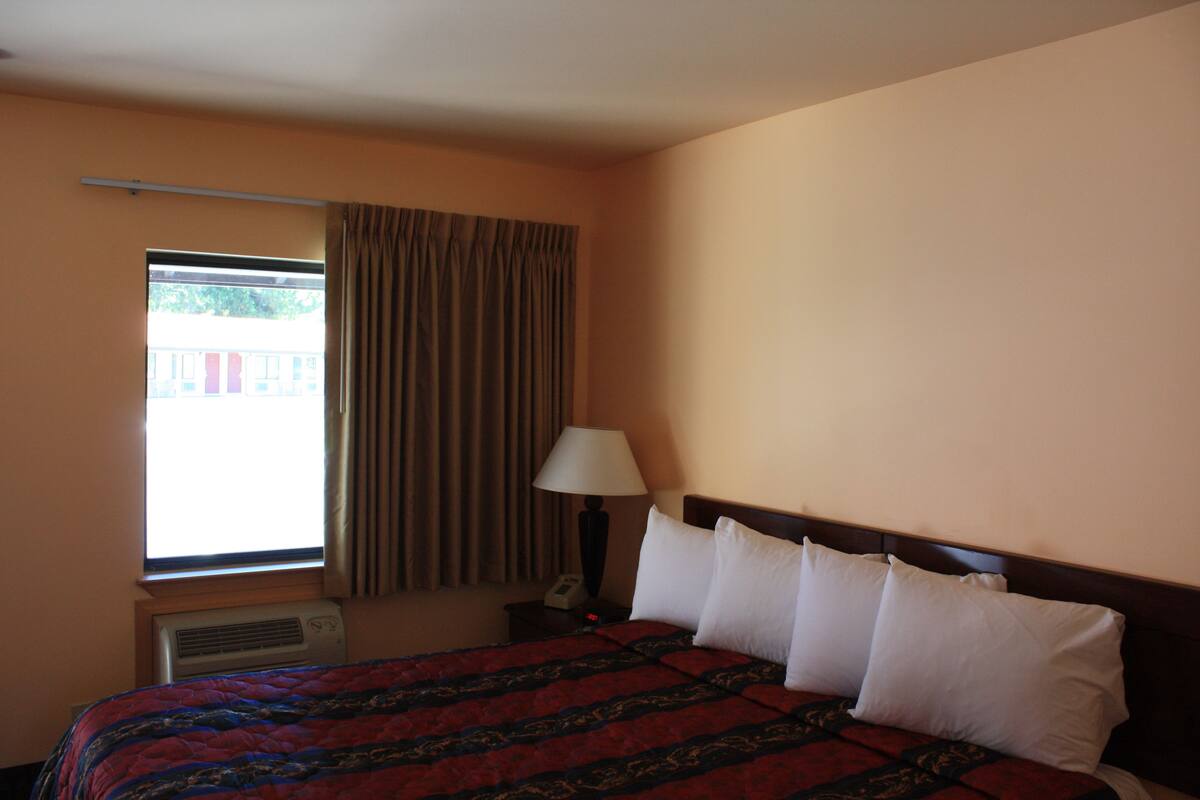 A hotel room with a bed, a window with curtains, an air conditioning unit, and a bedside table with a lamp.