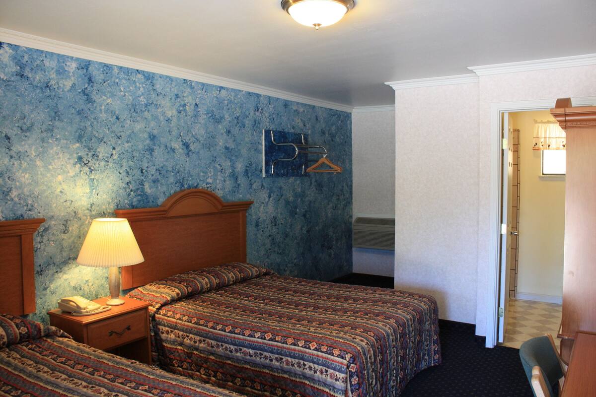 A hotel room with two beds, a nightstand with a lamp, a wooden headboard, a blue textured wall, a coat rack, and a door leading to another room.