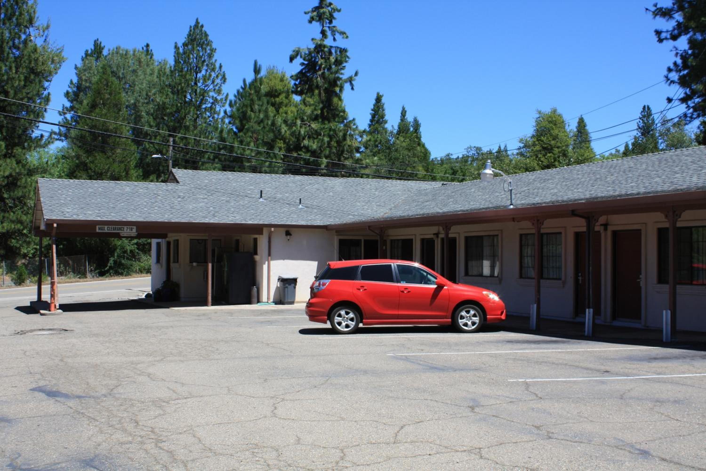 A motel with a red car parked in front, a covered parking area, and a building with a sign indicating 'Mail Clearance 7ft'.