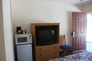 Pequeña habitación con un mueble de madera que contiene un televisor, un microondas y una cafetera. Hay una silla y una puerta con un letrero con un número.