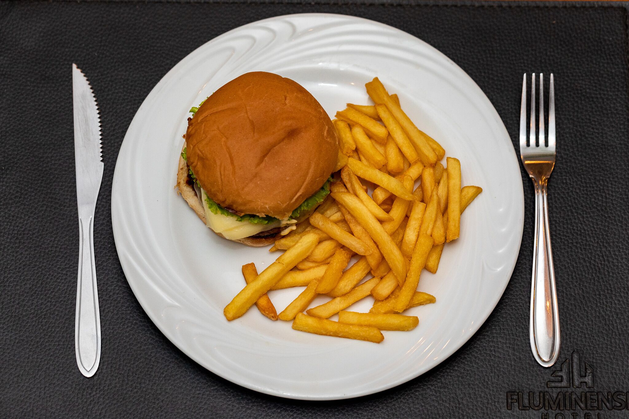 Un piatto con un hamburger e patatine fritte, su un piatto bianco, adagiato su una tovaglia scura.