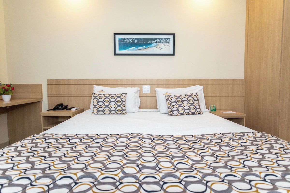 A hotel room with a large bed, two pillows with a geometric pattern, a framed picture on the wall, a bedside table with a phone, and a small plant.