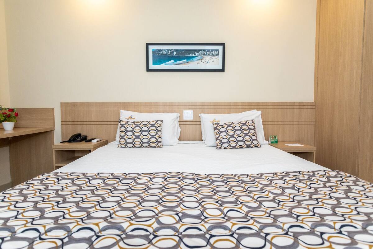 Ein Hotelzimmer mit einem großen Bett, zwei Kissen mit geometrischem Muster, einem gerahmten Bild an der Wand, einem Nachttisch mit Telefon und einer kleinen Pflanze.