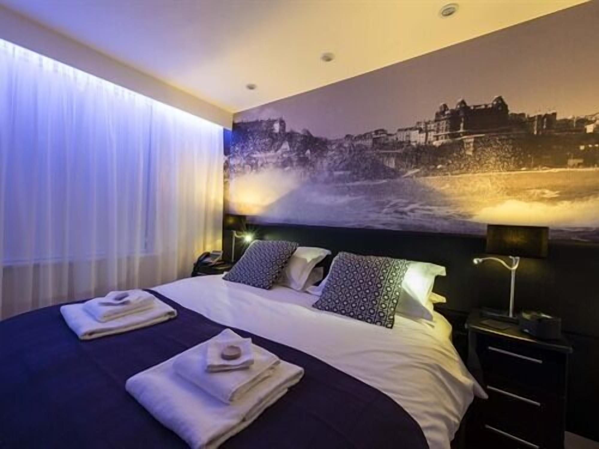 Une chambre d’hôtel avec un grand lit, deux oreillers, une table de chevet, une lampe et une fresque murale représentant un paysage côtier.