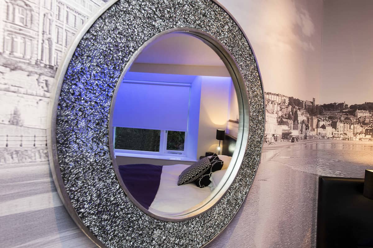 A bedroom with a large mirror, a bed, a window, and a cityscape mural on the wall.