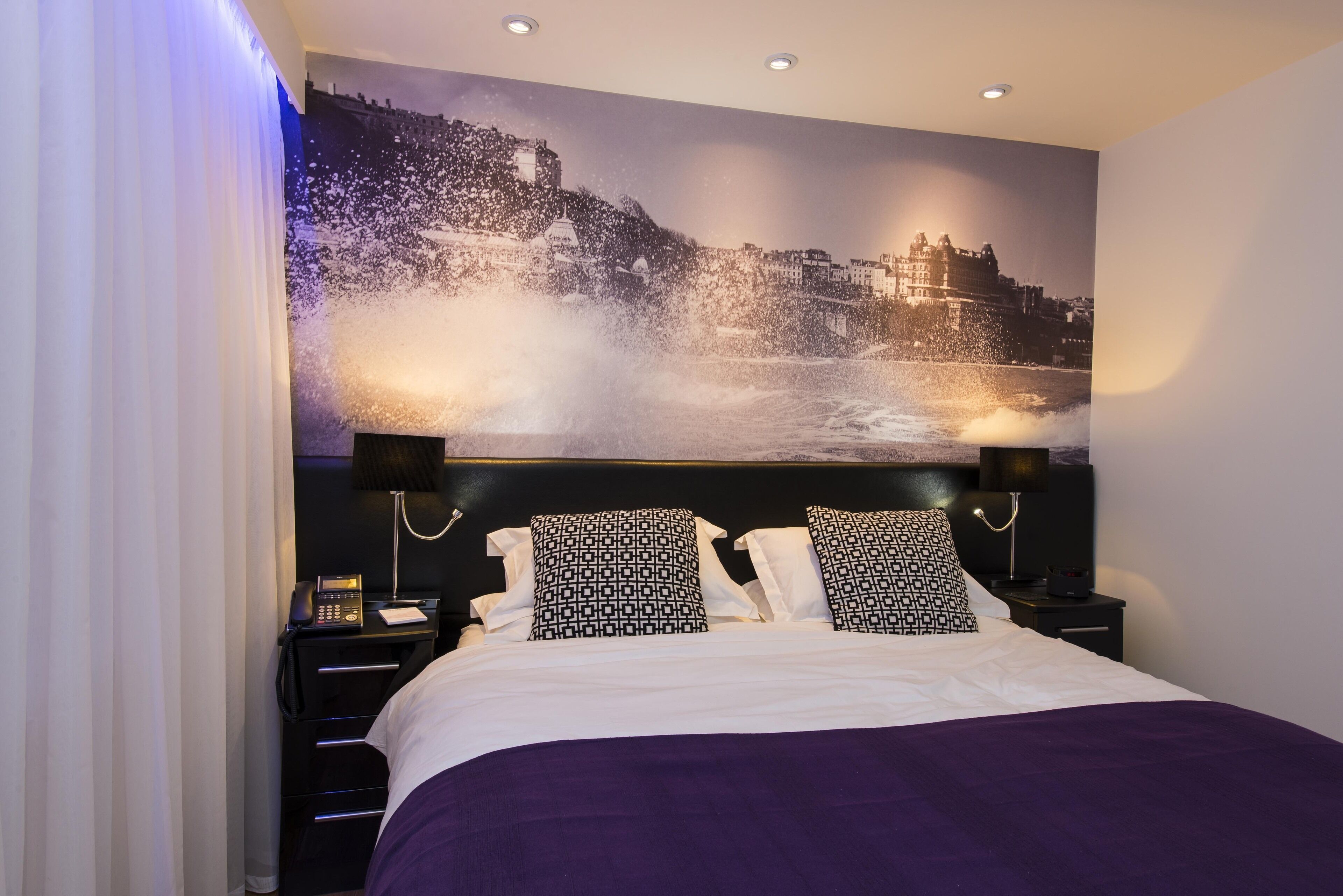 Une chambre d’hôtel avec un grand lit, deux tables de chevet, une petite table d’appoint, un téléphone fixé au mur et une grande fresque murale représentant un paysage.