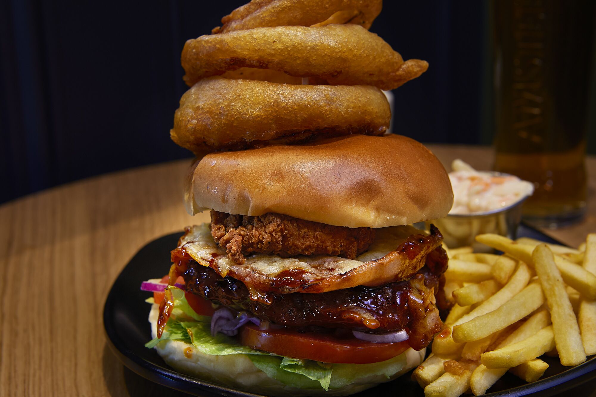 A burger with multiple layers of ingredients, including a fried chicken patty, cheese, lettuce, tomato, and onion, served with a side of French fries and a small bowl of sauce.