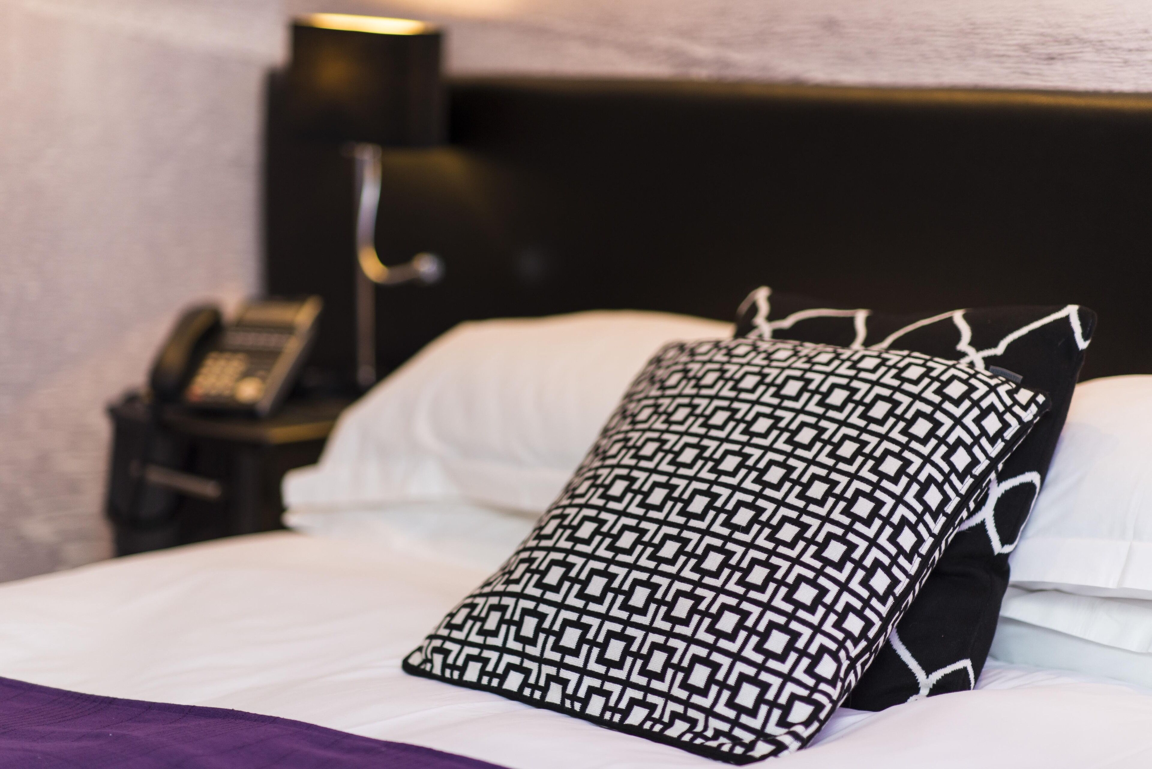 Una cama bien hecha con un cojín estampado, un teléfono en la mesita de noche y una lámpara de pared.