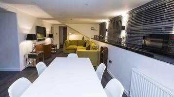 A modern hotel room with a dining table, white chairs, a sofa, and a kitchen area with a microwave.