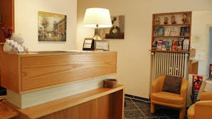 Una zona de recepción con un mostrador de madera, una lámpara, una silla, una estantería con libros y figuritas, y un cuadro en la pared.
