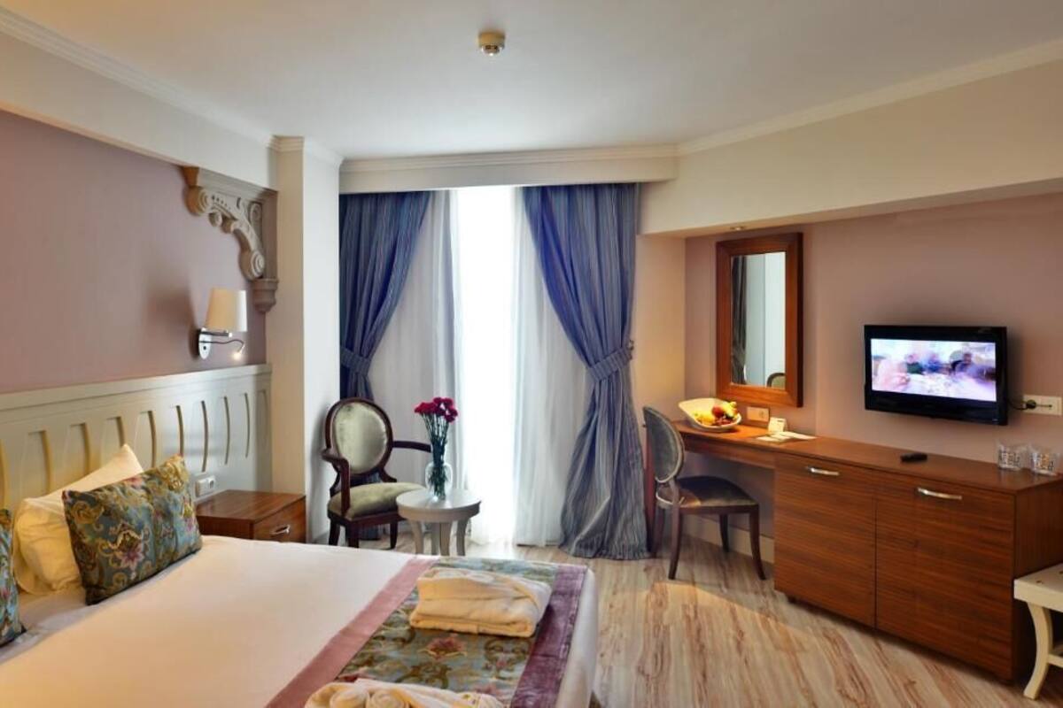 Habitación de hotel con cama, escritorio, silla, televisión y ventana con cortinas.