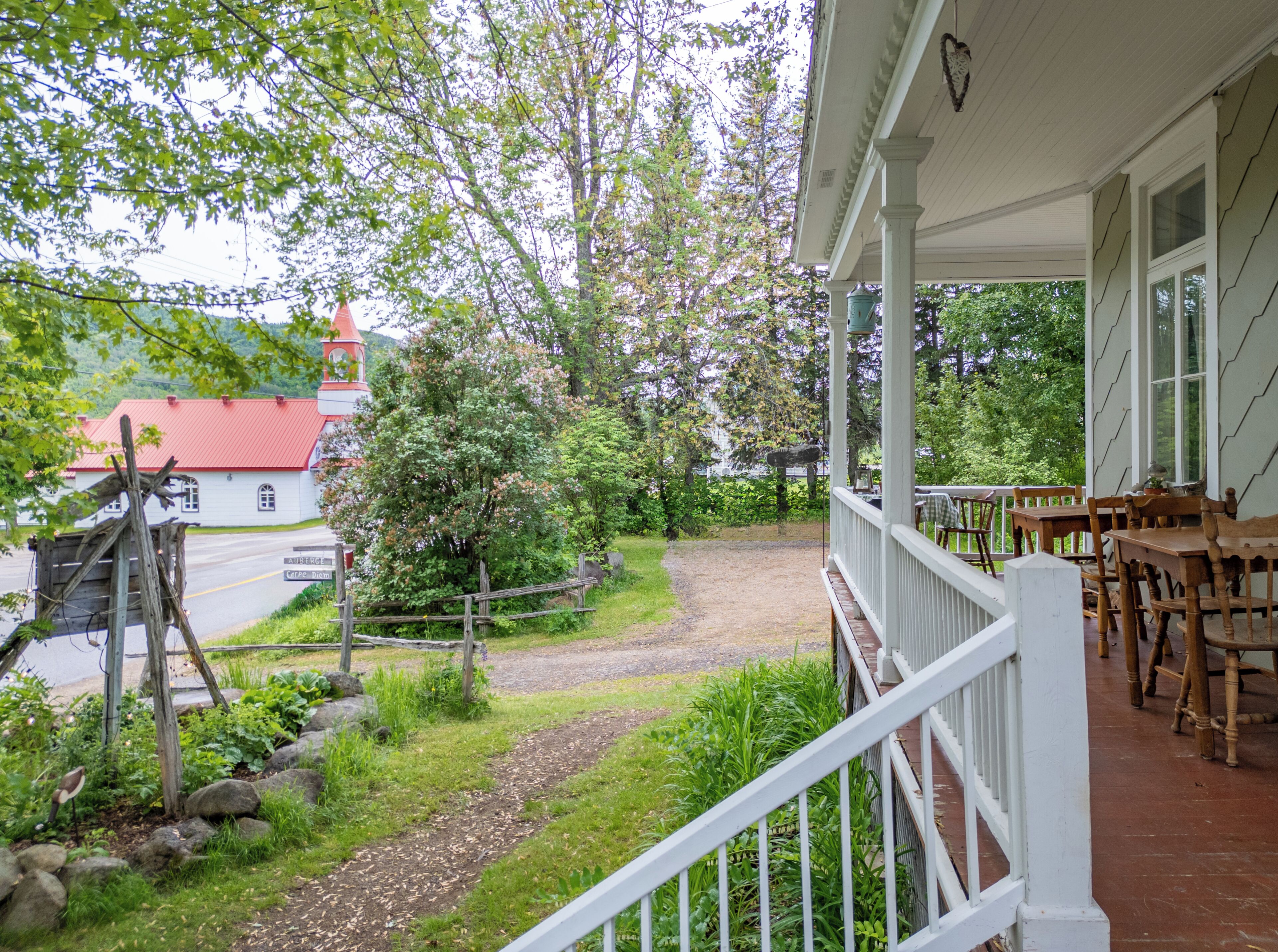 A covered porch with wooden furniture, a gravel path, and a view of a church with a red roof.