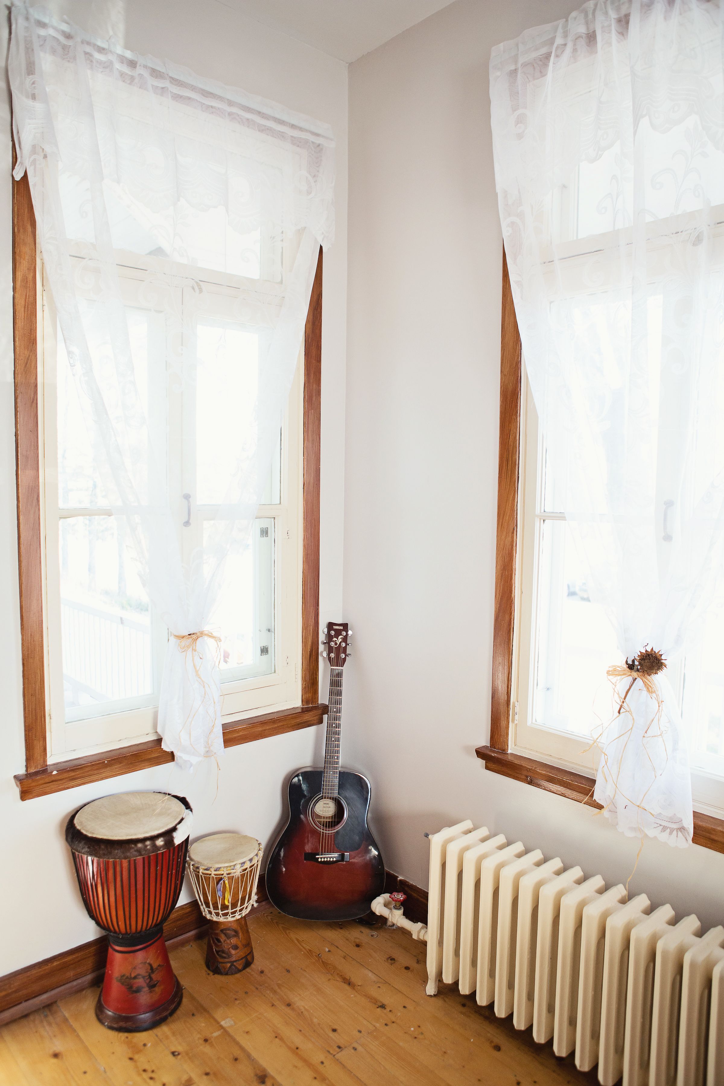Una habitación con dos ventanas, un radiador, una guitarra y dos tambores.