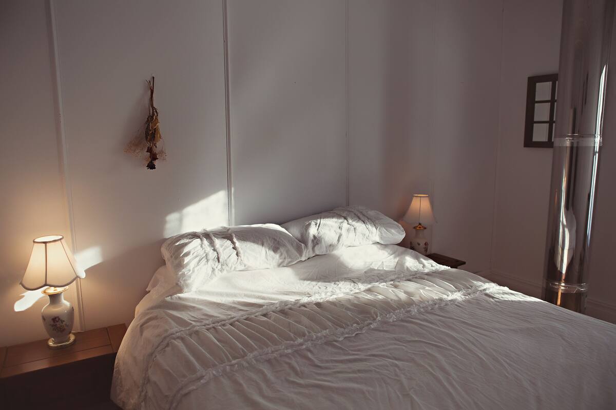 Ein ordentlich bezogenes Bett mit weißen Bettwäsche und einer mit Spitze verzierten Decke, zwei Nachttische mit Lampen sowie ein an der Wand befestigter Bilderrahmen.