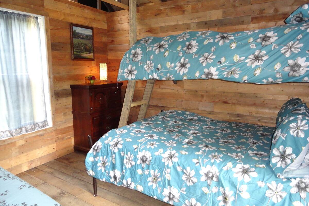 Ein Zimmer einer Holzhütte mit einem Etagenbett, einem Schminktisch, einer Leiter, einem Fenster mit Vorhängen und einem gerahmten Bild an der Wand.