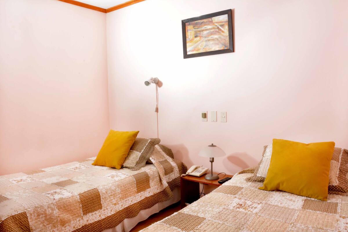Une chambre avec deux lits, chacun agrémenté d’un oreiller jaune, une table de chevet avec une lampe, un téléphone et un tableau encadré au mur.