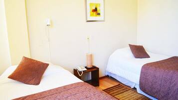 A hotel room with two beds, each with a brown pillow and a patterned bedspread. There is a small bedside table with a lamp and a phone. A framed picture hangs on the wall above the beds.