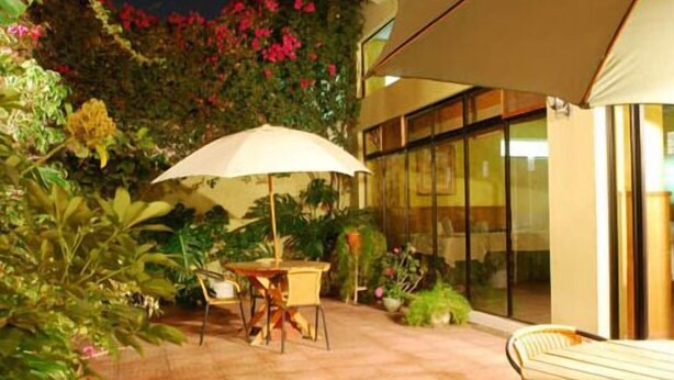Un patio al aire libre con mesa y sillas bajo un toldo, rodeado de abundante vegetación y un ventanal que da al interior.