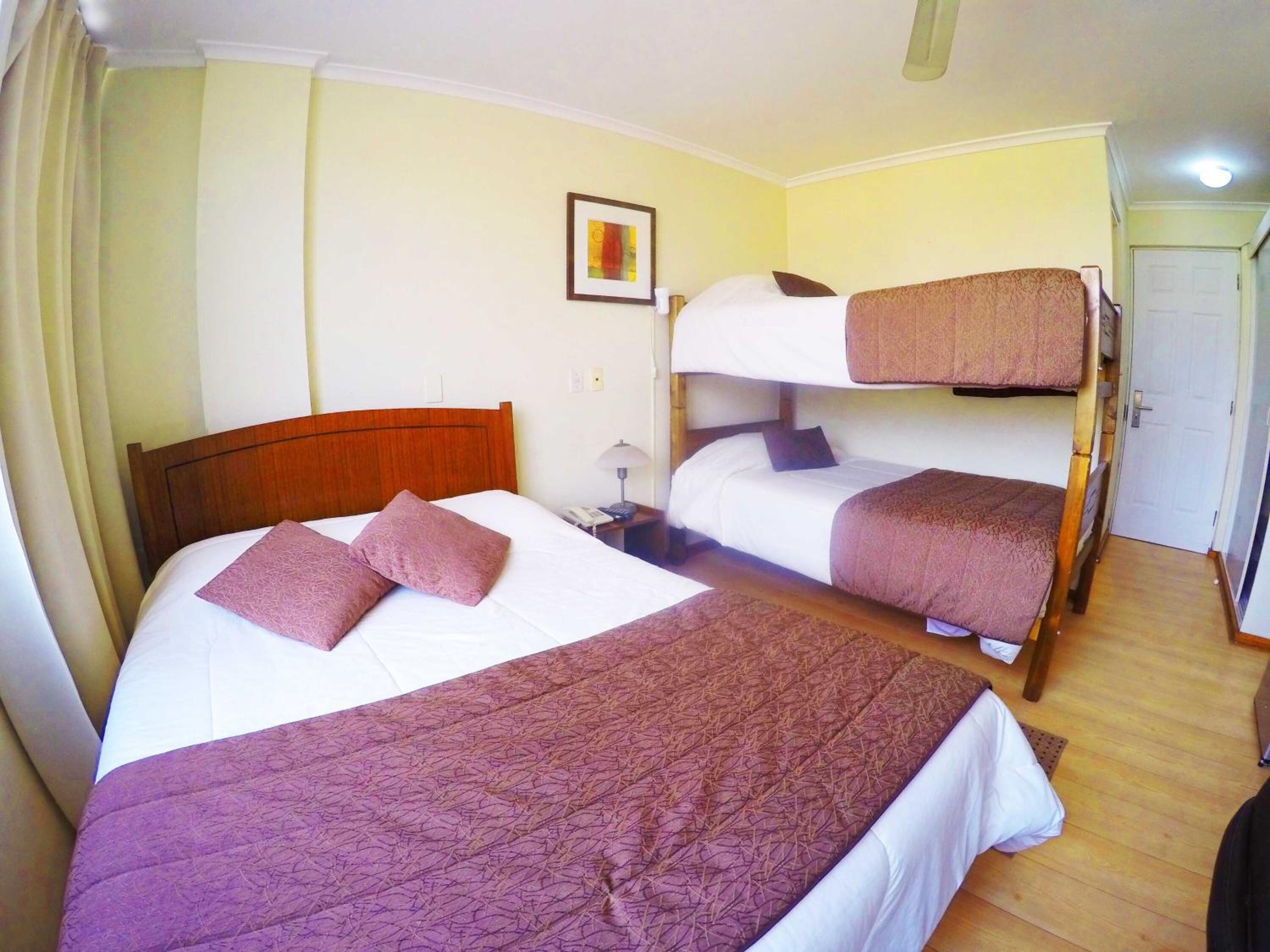 A hotel room with two beds, one with a wooden headboard and the other with a bunk bed. There is a bedside table with a lamp, a framed picture on the wall, and a door to the right.