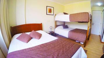 A hotel room with two beds, one with a wooden headboard and the other with a bunk bed. There is a bedside table with a lamp, a framed picture on the wall, and a door to the right.