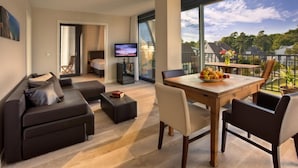A modern living room with a sofa, a dining table with chairs, a TV, and a balcony with a view of trees and houses. - Suite Hotel Binz Familienhotel Rügen (Binz)