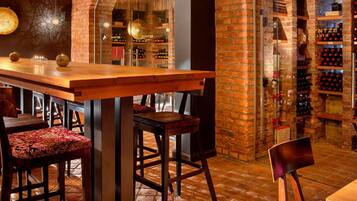 A bar area with wooden tables and chairs, a brick wall with wine racks, and a hanging light fixture.