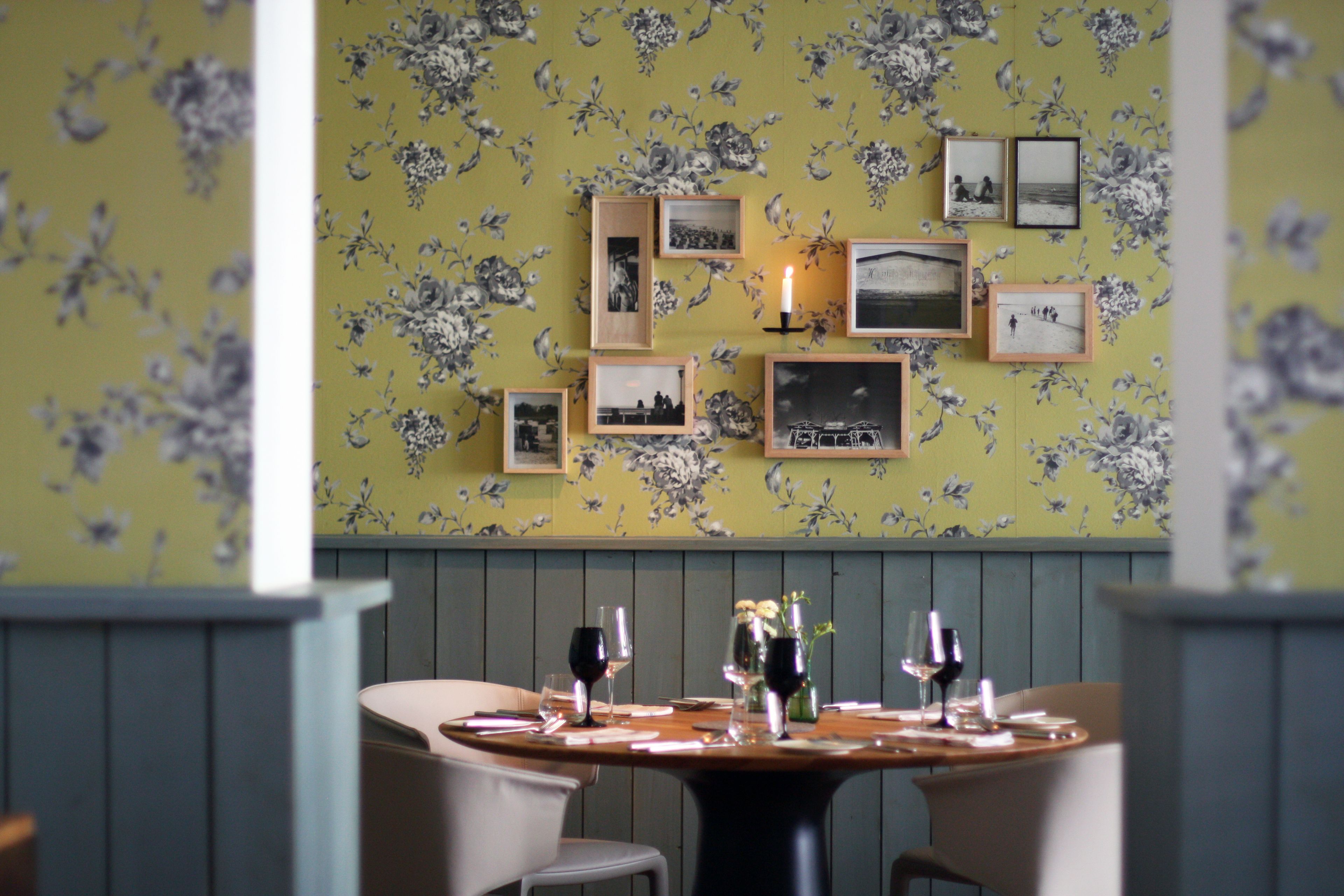 A dining area with a floral wallpaper, framed pictures on the wall, and a table set for two with wine glasses and a candle.