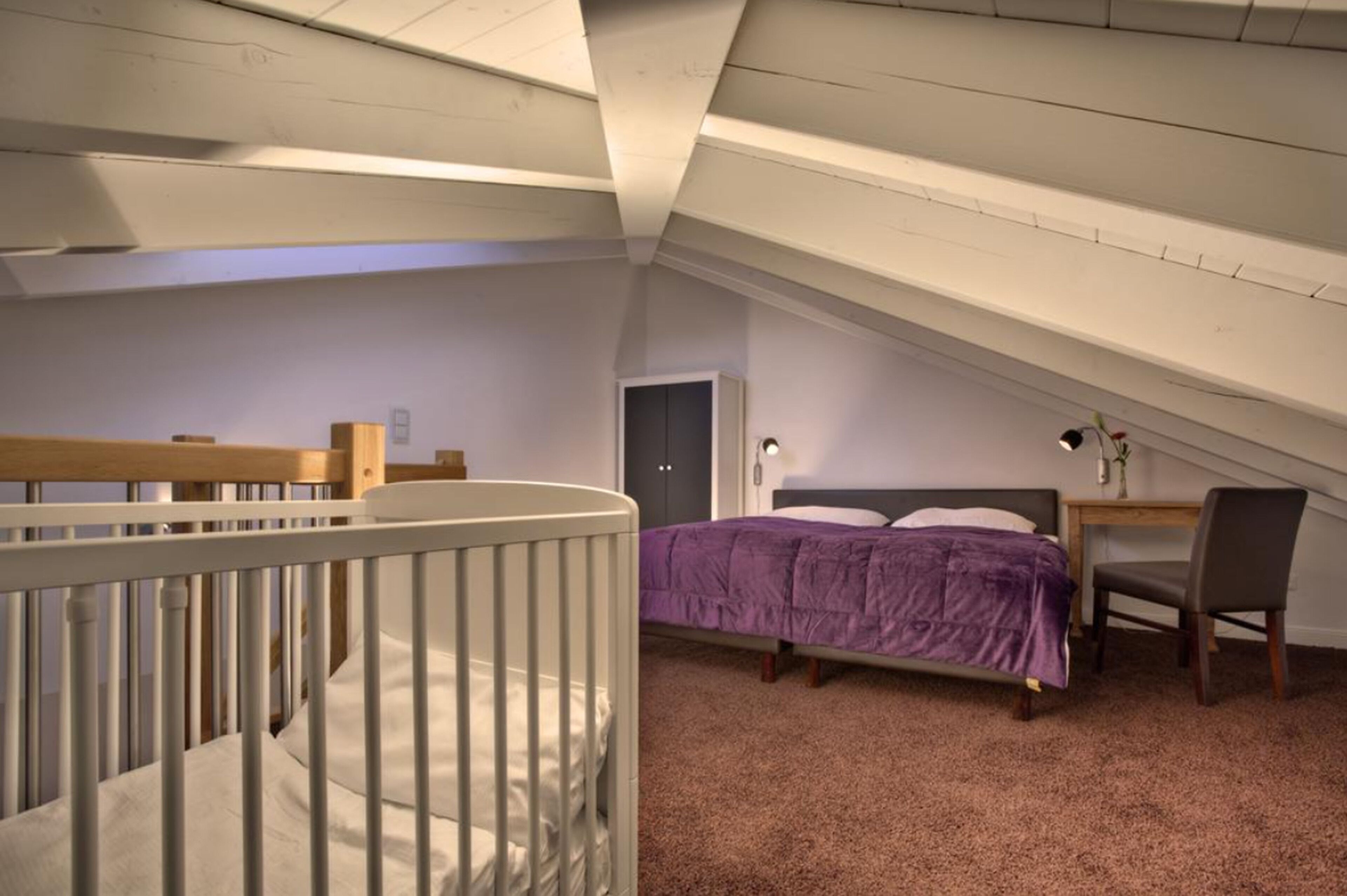A bedroom with a crib, a bed with a purple blanket, a desk with a chair, and a door.