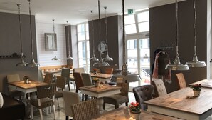 A restaurant with multiple tables and chairs, hanging pendant lights, and large windows allowing natural light. - Suite Hotel Binz Familienhotel Rügen (Binz)